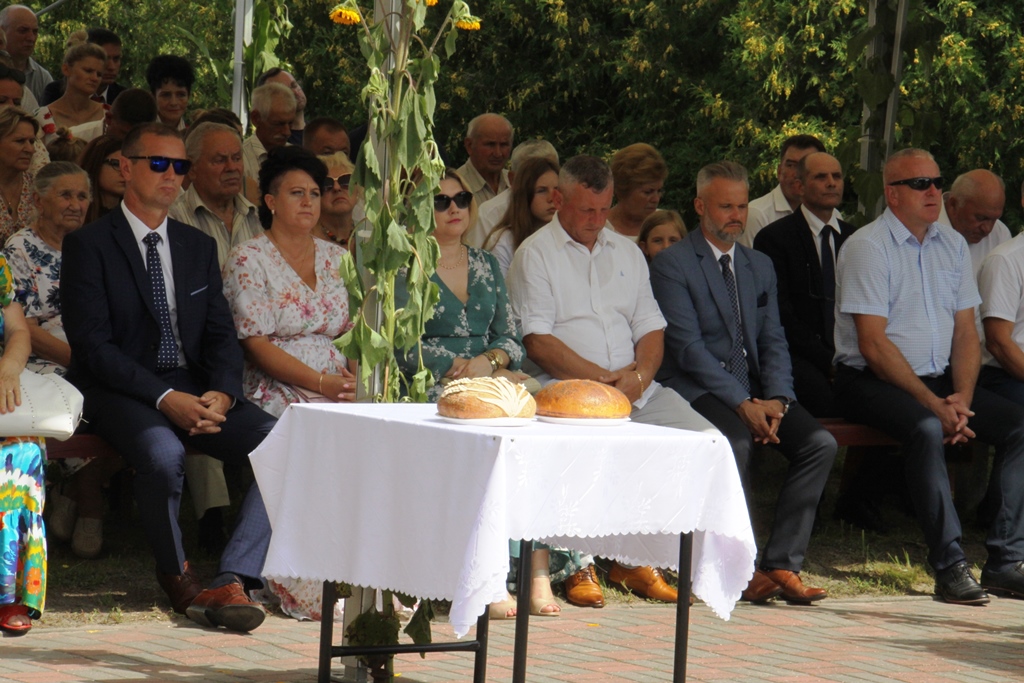 Uczestnicy dożynek siedzą na krzesłach w tym zaproszeni goście, na pierwszym planie dwa bochenki chleba